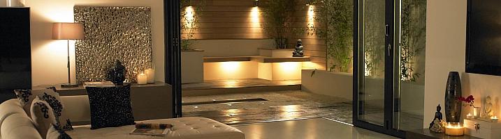 Modern living room at night with black open-corner bifold doors, leading out to a lit contemporary patio