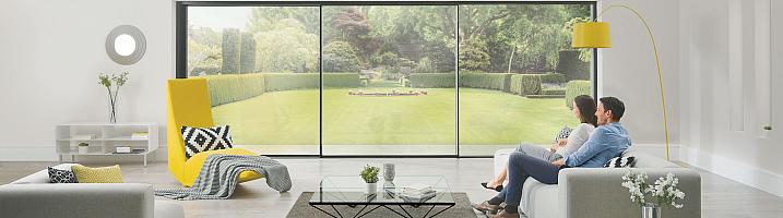 Two people sit on a sofa, looking out of a large, floor-to-ceiling slimline sliding door onto a manicured lawn.