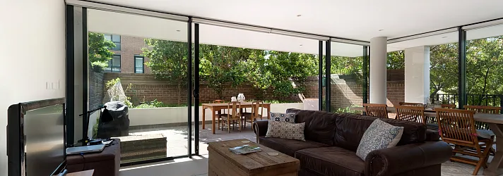 Bright living room with a brown leather sofa and large black sliding doors open to a sunny patio with a dining table