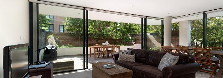 Bright living room with a brown leather sofa and large black sliding doors open to a sunny patio with a dining table