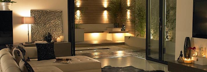 Modern living room at night with black open-corner bifold doors, leading out to a lit contemporary patio