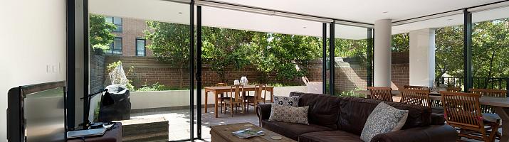 Bright living room with a brown leather sofa and large black sliding doors open to a sunny patio with a dining table