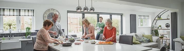 A family prepares food in a modern open-plan kitchen, with a large, dark grey bifolding door in the background