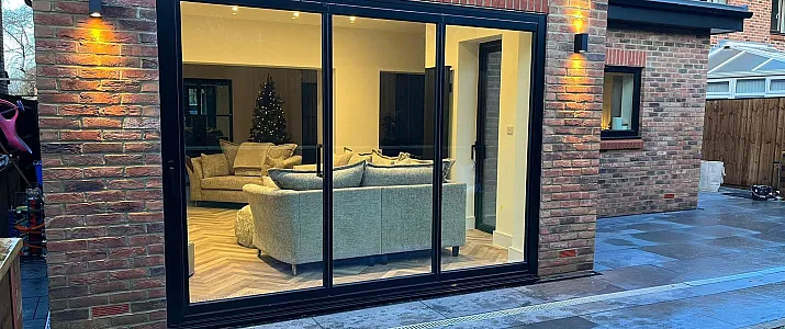 Black three-panel sliding door on a brick house at night, looking into a lit living room with sofas and a Christmas tree