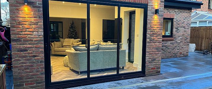 Black three-panel sliding door on a brick house at night, looking into a lit living room with sofas and a Christmas tree