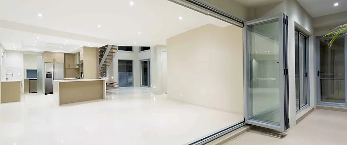 Silver aluminum bifold doors fully folded open, revealing a large, modern, empty room with a kitchen and tiled floors