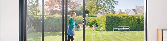 A person opening a black-framed slim sliding door, leading out to a sunny garden with a bench