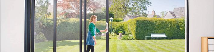 A person opening a black-framed slim sliding door, leading out to a sunny garden with a bench