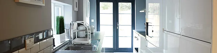 A modern galley kitchen with white gloss cabinets and dark grey double doors at the far end leading to a patio