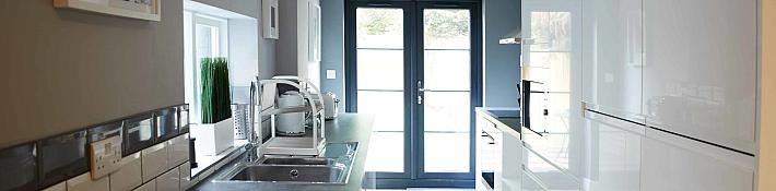 A modern galley kitchen with white gloss cabinets and dark grey double doors at the far end leading to a patio