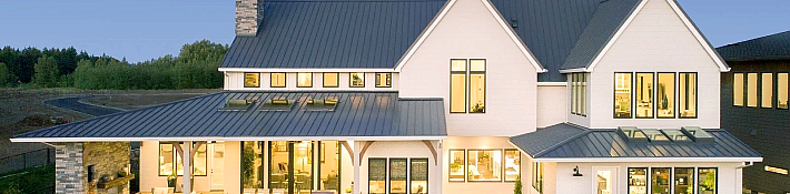 The rear exterior of a large, white modern farmhouse at dusk, featuring a metal roof, a large lawn, and a lit covered patio
