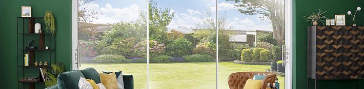 A modern living room with a green sofa and a large, white-framed sliding door providing a view of a lush garden.