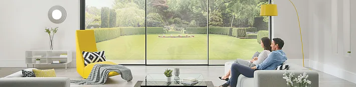 Two people sit on a sofa, looking out of a large, floor-to-ceiling slimline sliding door onto a manicured lawn.