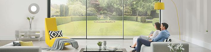Two people sit on a sofa, looking out of a large, floor-to-ceiling slimline sliding door onto a manicured lawn.