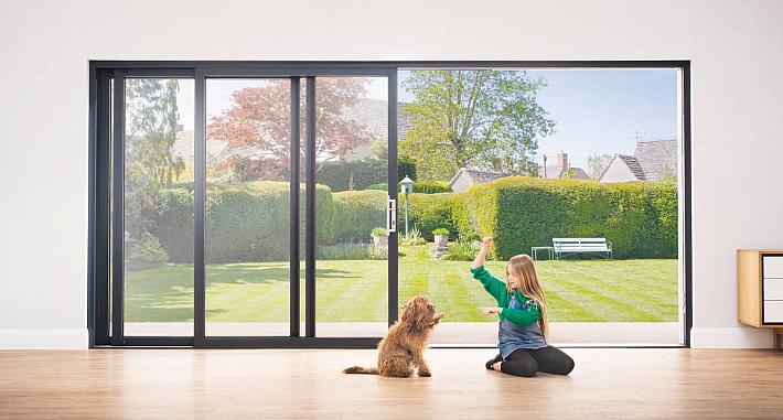 A girl plays with a dog on a wood floor in front of a large, black Origin sliding door open to a sunny garden