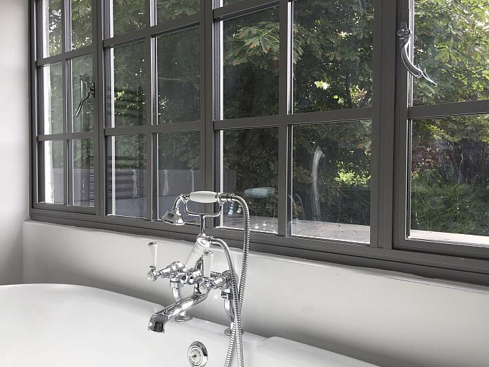 Close-up of a grey Aluco steel-look window with a grid pattern, set above a freestanding bathtub with classic chrome taps