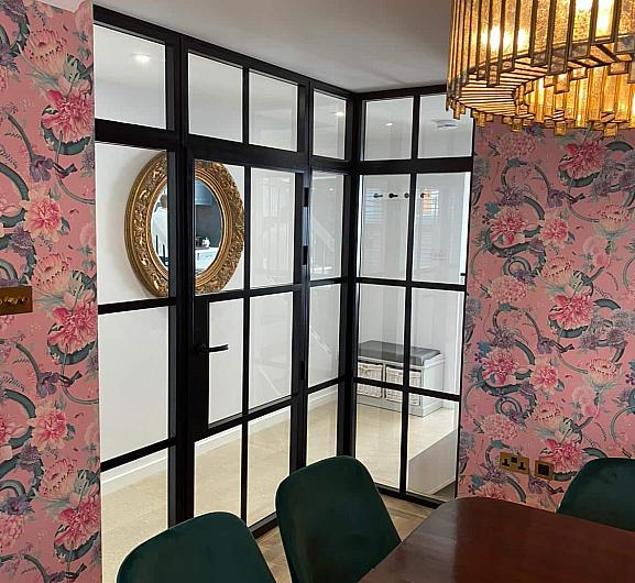 Internal black steel-look corner screen and door in a dining room with pink floral wallpaper and green velvet chairs