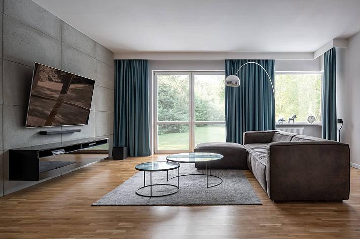 Modern living room with a brown sofa, concrete wall, and a large sliding glass door with blue curtains, looking onto a garden