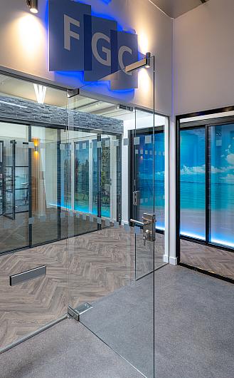 Entrance to the FGC showroom, showing frameless glass doors, a blue backlit FGC logo, and herringbone floors inside