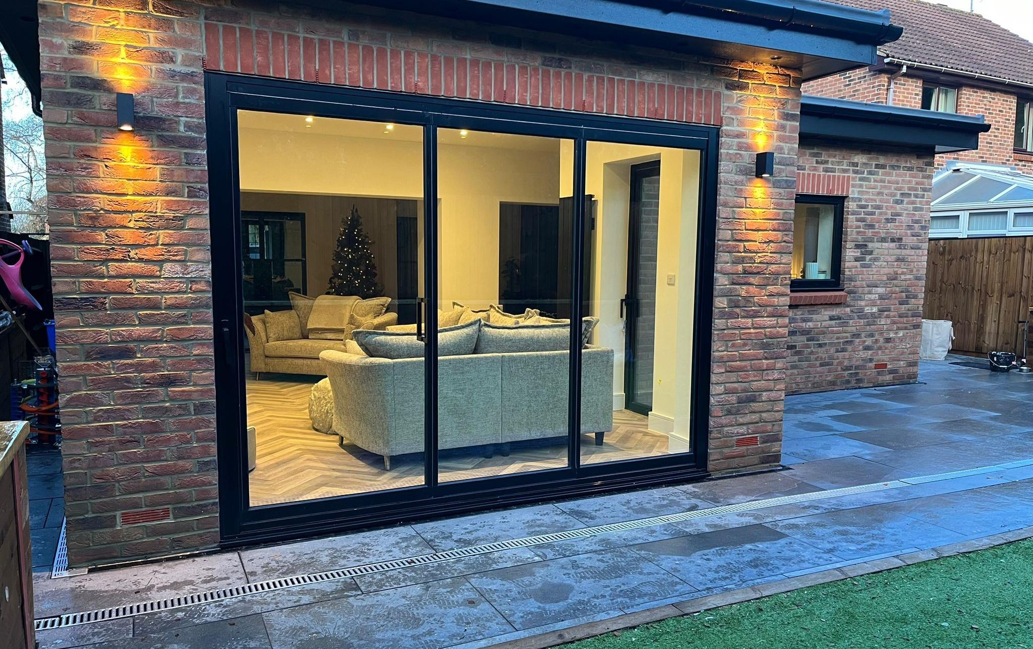 Black three-panel sliding door on a brick house at night, looking into a lit living room with sofas and a Christmas tree