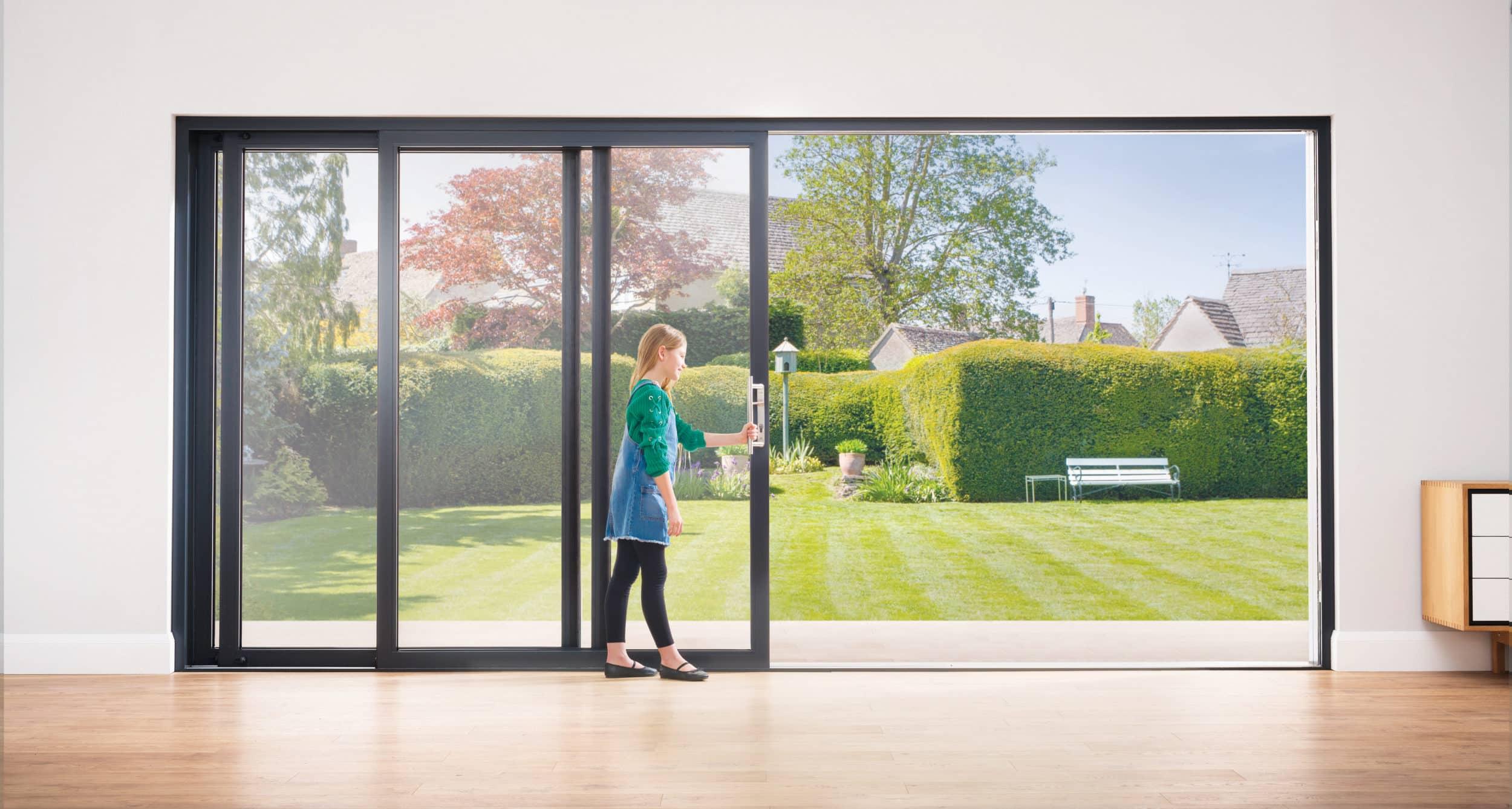 A person opening a black-framed slim sliding door, leading out to a sunny garden with a bench