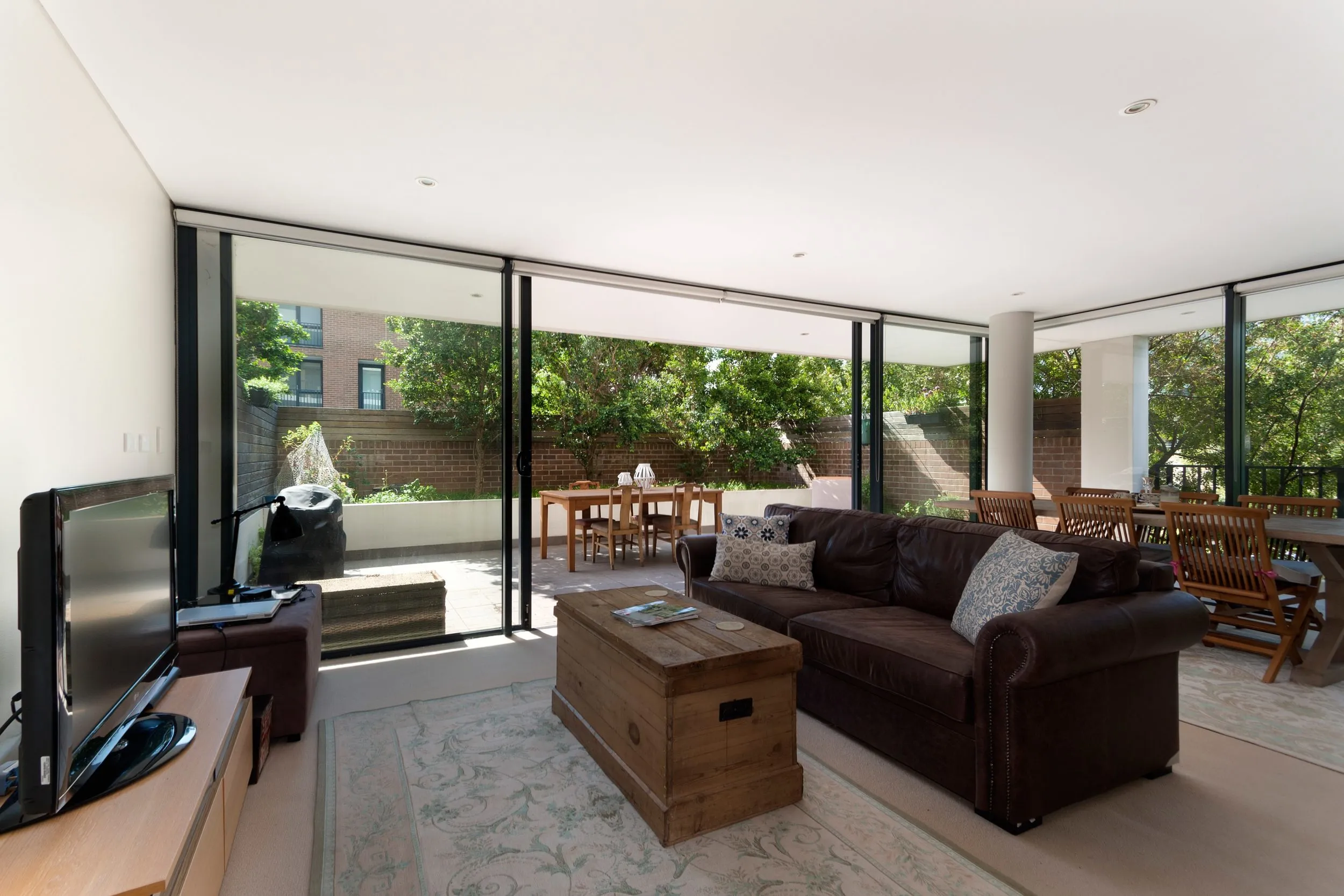 Bright living room with a brown leather sofa and large black sliding doors open to a sunny patio with a dining table