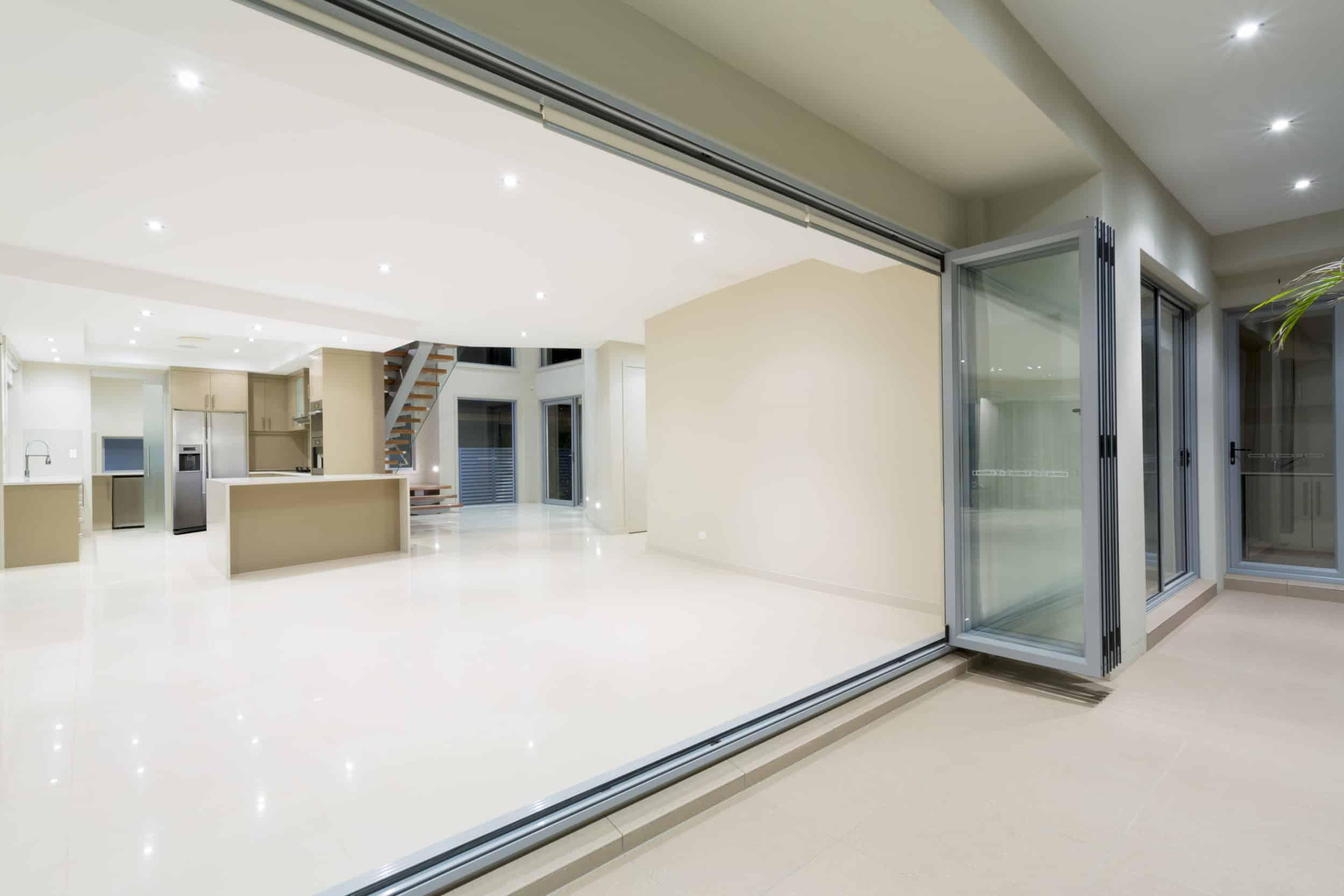 Silver aluminum bifold doors fully folded open, revealing a large, modern, empty room with a kitchen and tiled floors