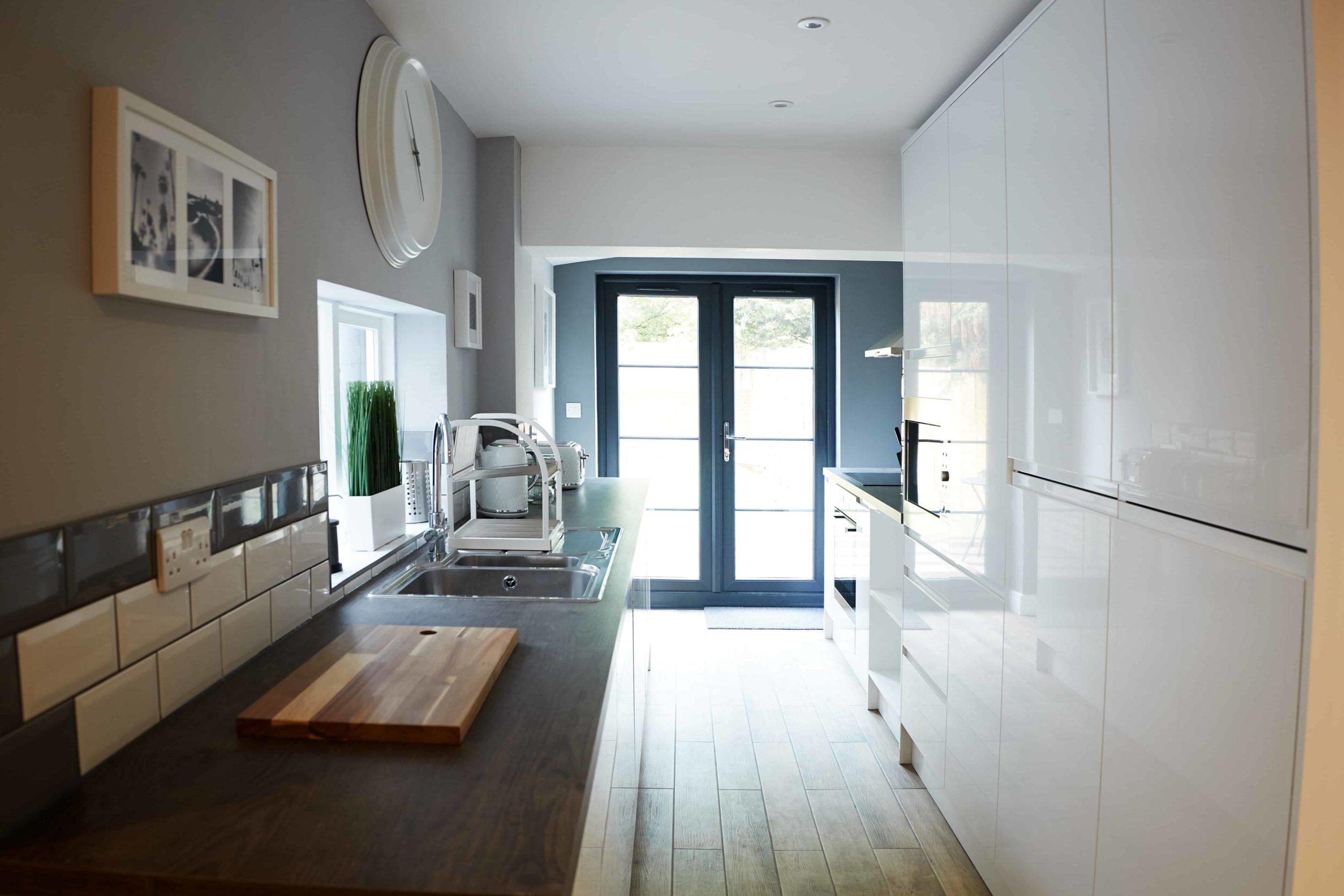 A modern galley kitchen with white gloss cabinets and dark grey double doors at the far end leading to a patio