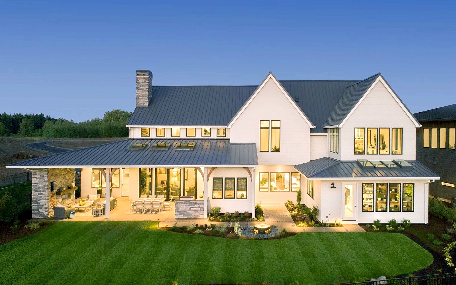 The rear exterior of a large, white modern farmhouse at dusk, featuring a metal roof, a large lawn, and a lit covered patio