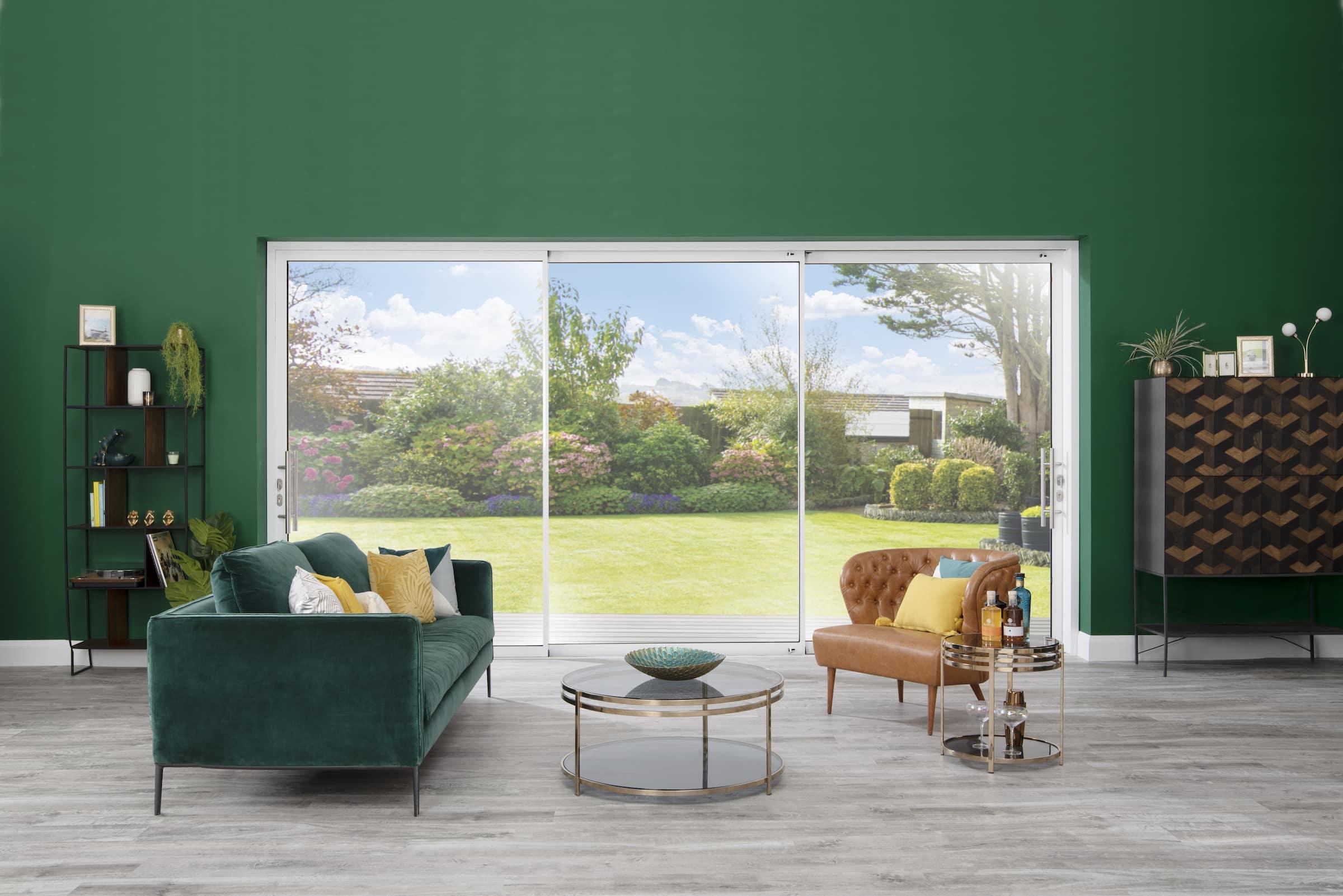 A modern living room with a green sofa and a large, white-framed sliding door providing a view of a lush garden.