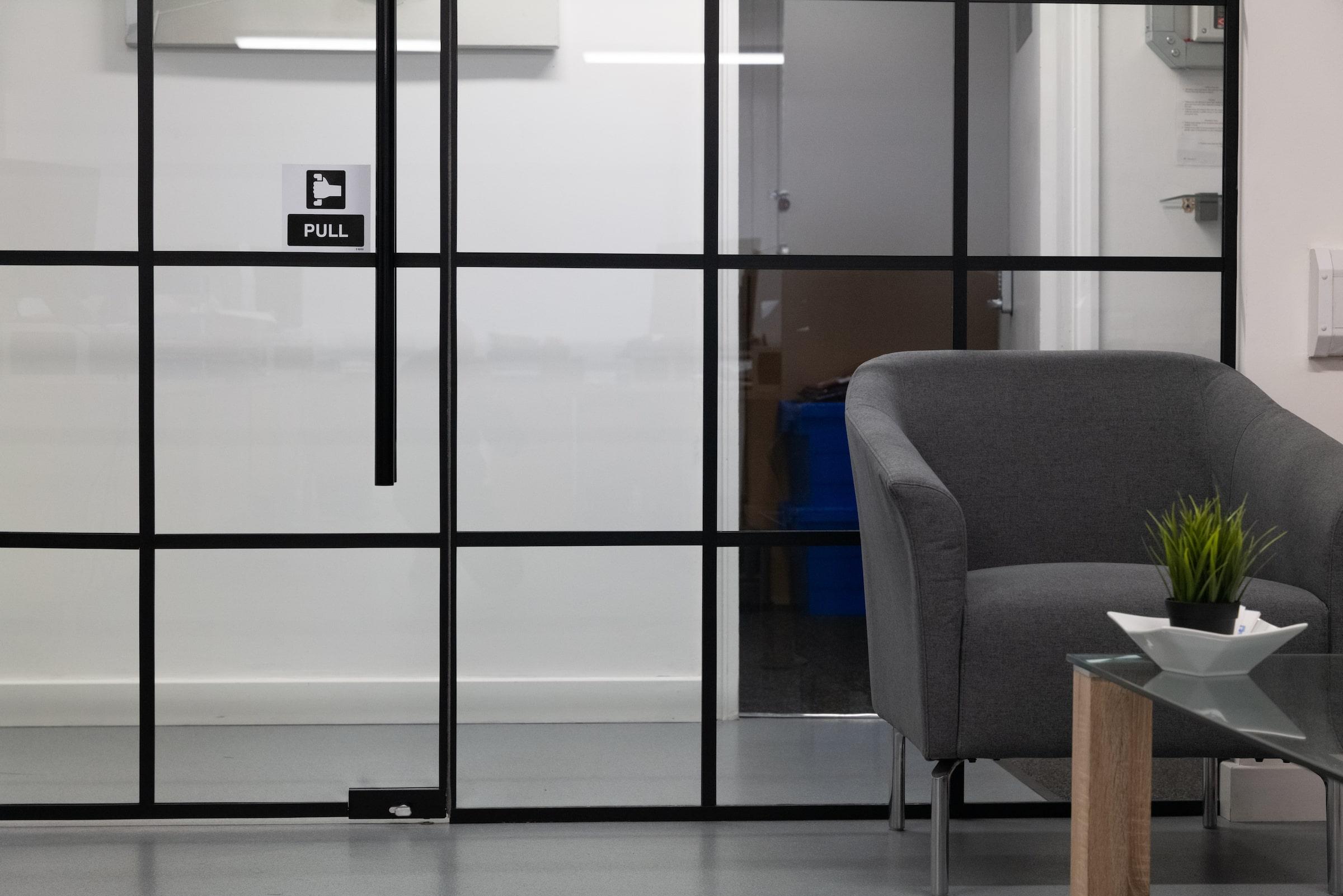 A long black steel-look glass screen and door in an office corridor, with a 'PULL' sticker and a large plant leaf in the foreground