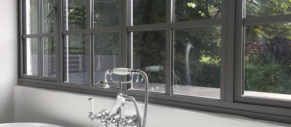 Close-up of a grey Aluco steel-look window with a grid pattern, set above a freestanding bathtub with classic chrome taps