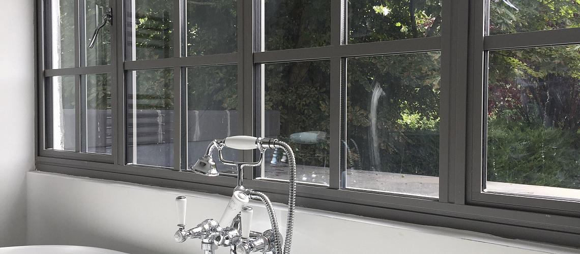 Close-up of a grey Aluco steel-look window with a grid pattern, set above a freestanding bathtub with classic chrome taps