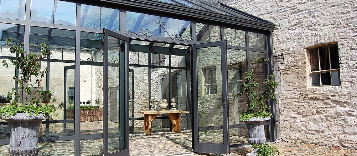 Exterior of a grey Alitherm glass conservatory with an open double door, set against a rustic stone building