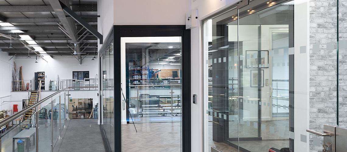 Modern showroom hallway with a glass balustrade, overlooking a factory. Black-framed and frameless glass doors are on the right