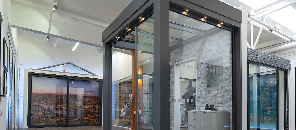 A bright showroom with herringbone floors, displaying a modern grey glass garden room and other door products