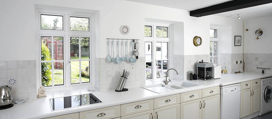 A white kitchen with cream cabinets and three white Smart Systems windows with a grid pattern, looking onto a garden