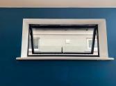 A black, top-hung heritage aluminium window, shown open, set in a deep white reveal against a dark blue wall.