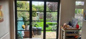 Exterior view of closed dark grey steel-look bifold doors set in a brick wall, showing reflections on the glass