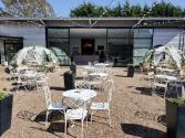 A sunny patio with white tables and dining igloos. The building's black steel-look sliding doors are wide open
