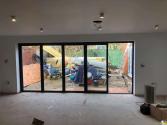 Five-panel black bifold door with the left-hand traffic door slightly open, viewed from inside an unfinished room