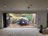Black bifold doors fully folded open, showing a wide opening from the unfinished room to the construction-filled garden