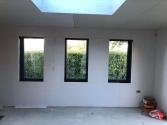 Three tall, narrow, dark grey windows installed in an unfinished drywall, looking out onto a tall green hedge
