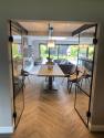 Fully open black steel-look double doors revealing a bright, open-plan dining and living area with herringbone floors.