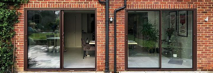 Exterior view of a red brick house with two sets of dark brown slimline sliding doors leading onto a grey paved patio.