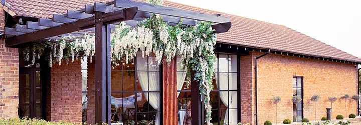 Exterior of a brick venue with a black, flower-draped pergola. Large, black steel-look windows and doors are set into the wall