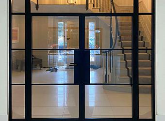 Closed black steel-look double doors with side panels, viewed from a hallway, looking into another hall with a staircase