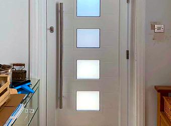 Closed white aluminum front door with a long vertical handle and four central, square frosted glass windows, viewed from inside