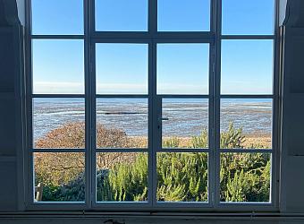 Interior view of a large white steel-look window, looking out over a sunny coastal landscape at low tide.