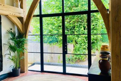 Rustic room with exposed beams, looking through a closed black steel-replacement French door onto a green garden